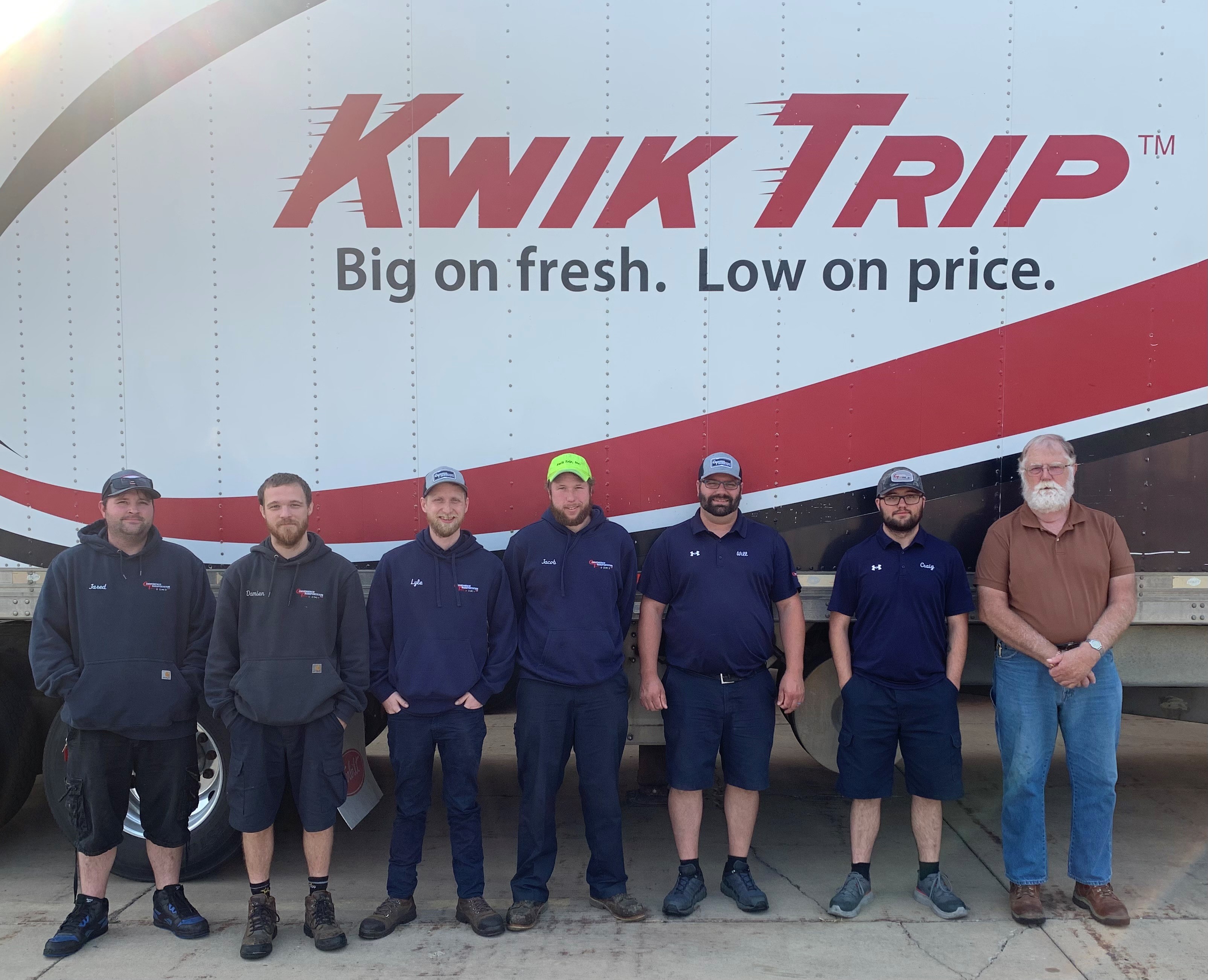 Six Kwik Trip employees completing the commercial driver's license for truck driving stand in front of a trailer that reads Kwik Trip, with their instructor, Val Gunmundson standing alongside them at the far right.